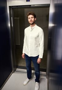 Witte button-up shirt met een Mandarin kraag, donkerblauwe jeans en witte sneakers, staand in een moderne lift met metalen muren.