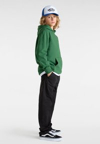Felpa verde con tasca frontale, abbinata a pantaloni neri e sneakers bianco-neri. Cappellino bianco e blu con un logo.