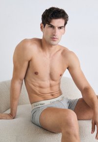 Huishoudelijke man zonder shirt met kort, krullend haar, zittend op een textuur witte stoel, gekleed in grijze Calvin Klein boxershorts, kijkend iets naar rechts.
