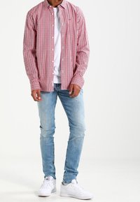 Camisa de cuadros rojos y blancos con botones, llevada sobre una camiseta blanca, combinada con jeans azul claro y zapatillas blancas.