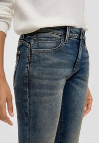 Niebieskie dżinsy z dopasowanym krojem, z wyblakłymi detalami, subtelnym efektem przetarć i kontrastowym szwem. Kieszenie zawierają ozdobne elementy metalowe.