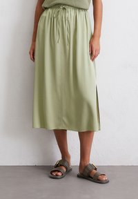 Elastic waist midi skirt in light green with a side slit and smooth texture, paired with dark sandals featuring a gold accent.