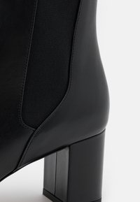 Gros plan d’une botte chevillère en cuir noir avec panneau latéral élastique et talon bloc épais sur fond blanc.