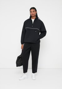 Giacca nera a pullover con colletto alto e una striscia orizzontale bianca sul petto, abbinata a pantaloni neri e sneakers bianche.