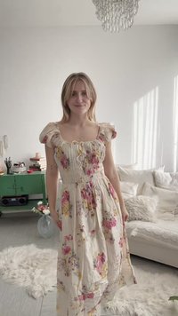 Fiatal nő virágmintás, fodros ujjú ruhában, aki egy világos nappaliban áll, ahol fehér bútorok, zöld szekrény és kristálycsillár található.