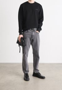 Mężczyzna w czarnej koszuli z długim rękawem, szarych dżinsach, czarnym pasku, czarnych butach, trzymający czarną, małą torbę, stoi na betonowej podłodze.