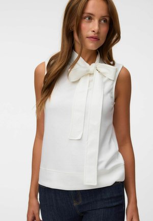 Femme portant un haut blanc sans manches avec un grand nœud papillon au cou, associé à un jean bleu foncé, debout devant un fond uni.
