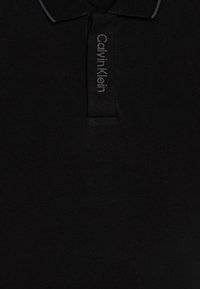 Polo noir en tissu texturé, avec un design à col et le logo "Calvin Klein" en gris foncé sur la patte de boutonnage.