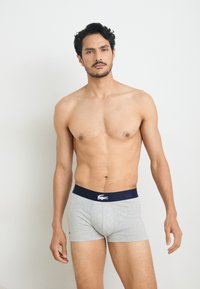 Grijze boxershorts met een marineblauwe tailleband met een wit logo. De stof lijkt zacht met een gladde textuur. Het model staat rechtop.