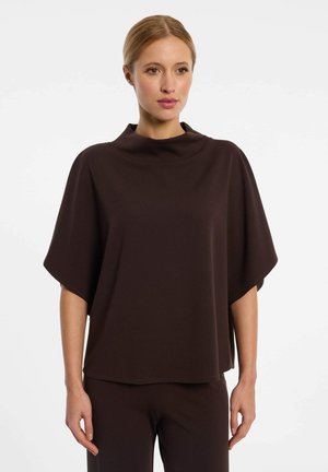 Vrouw met blond haar die een losse, donkerbruine top met korte mouwen en bijpassende broek draagt tegen een effen lichte achtergrond.