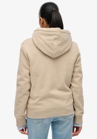 Beige hoodie van zacht, gestructureerd materiaal. Heeft een grote capuchon, geribbelde manchetten en zoom. Draagt over lichtblauwe spijkerbroek met zichtbare stiksels.