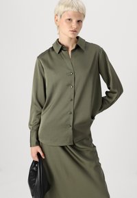 Chemise olive à manches longues avec col, fermeture à boutons sur le devant, texture lisse et coupe décontractée, associée à une jupe assortie et un sac noir.