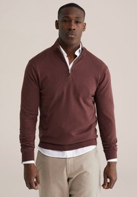 Pull léger bordeaux avec un col à demi-fermeture éclair, des poignets et un ourlet côtelés, porté avec une chemise blanche à col en dessous, associé à un pantalon beige.