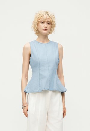 Femme aux cheveux blonds bouclés portant un haut sans manches bleu clair à peplum et un pantalon blanc ample, debout devant un fond clair uni.