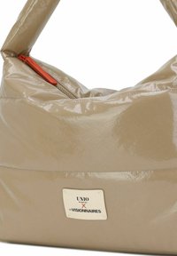 Bolso de piel sintética beige con forma curva, con una cremallera naranja y una etiqueta blanca con texto negro. Textura suave y brillante.