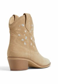 Botín de ante marrón con bordados florales, tacón afilado y costuras detalladas. Presenta una puntera puntiaguda y un diseño texturizado en el tacón.