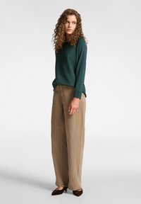 Top verde de manga larga con un ajuste relajado, combinado con pantalones de pierna ancha beige. Zapatos de terciopelo marrón completan el conjunto.