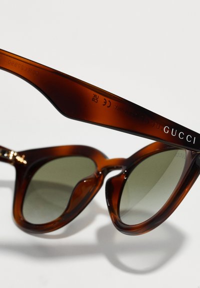 Smeđe kornjačaste sunčane naočale s kutnim, prevelikim okvirom. Tamnozelena sočiva i srebrni detalji s otisnutim "GUCCI" na drškama.