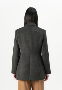 Femme aux longs cheveux noirs, portant un blazer ajusté gris foncé et un pantalon marron, se tenant face à un fond blanc avec le dos tourné vers le spectateur.