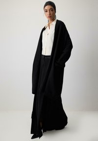 Melna pārmērīga izmēra kardigāna virs baltas pogu krekla, apvienota ar melnām svārkiem. Gluda tekstūra un minimālistisks dizains ar sānu šķēlumu.