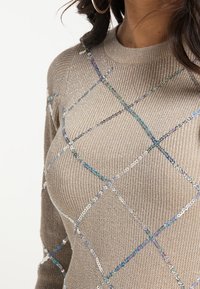 Beige stickad tröja med ett diamantsmönster gjort av paljetter i blå, silver och iriserande färger, med rund halsringning och långa ärmar.