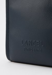 Sac en cuir marine avec une texture granuleuse et un logo embossé "LANCEL PARIS 1876". Forme structurée avec des détails de couture contrastés.