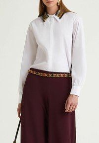 White shirt with decorative gold and silver embellishments on the collar, paired with burgundy wide-leg trousers and a multicolored chain belt.