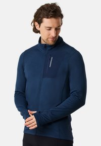 Marineblaue Zip-Jacke aus strukturiertem Material, mit hohem Kragen und taillierter Passform. Sie verfügt über eine Brusttasche mit sichtbarem Logo. Daumenlöcher an den Ärmeln.