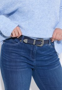 Persona vistiendo un suéter de punto azul ajustando un cinturón negro con hebilla de plata grabada sobre unos jeans de color azul oscuro.