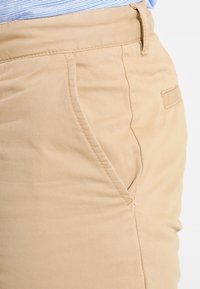 Pantalon en coton beige avec une coupe ajustée, des poches frontales diagonales et une texture lisse avec des détails de couture subtils.