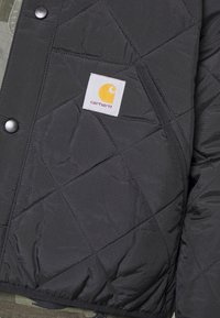 Zwarte gewatteerde jas met diagonale stiksels, drukknoopsluiting en een Carhartt-logo patch. Voorzien van een ritszak voor extra functionaliteit.