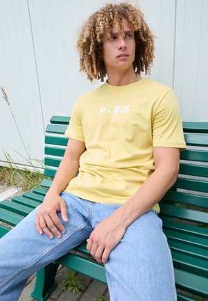 T-shirt di cotone gialla con testo "PARIS" in bianco, abbinata a jeans di denim blu chiaro, indossata mentre è seduta su una panchina verde.