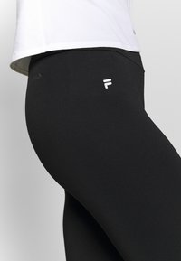 Schwarze Sportleggins aus einem glatten, dehnbaren Material, mit einem weißen Logo auf der Vorderseite und einem hoch sitzenden Design.
