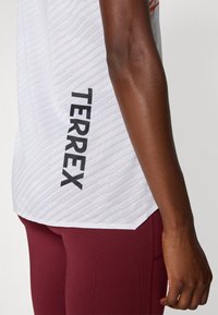 Haut de sport blanc avec un motif texturé et "TERREX" imprimé en noir ; associé à des leggings bordeaux, dotés d'un tissu lisse et d'une coupe ajustée.