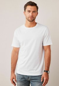 T-shirt en coton blanc à manches courtes, col rond et texture lisse. Porté avec un jean bleu et une montre noire. Design simple.