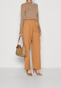 Beiger Pullover mit asymmetrischem Design und goldenen Knöpfen, kombiniert mit hoch taillierten, weit geschnittenen Karamellhosen und einer gemusterten Handtasche.