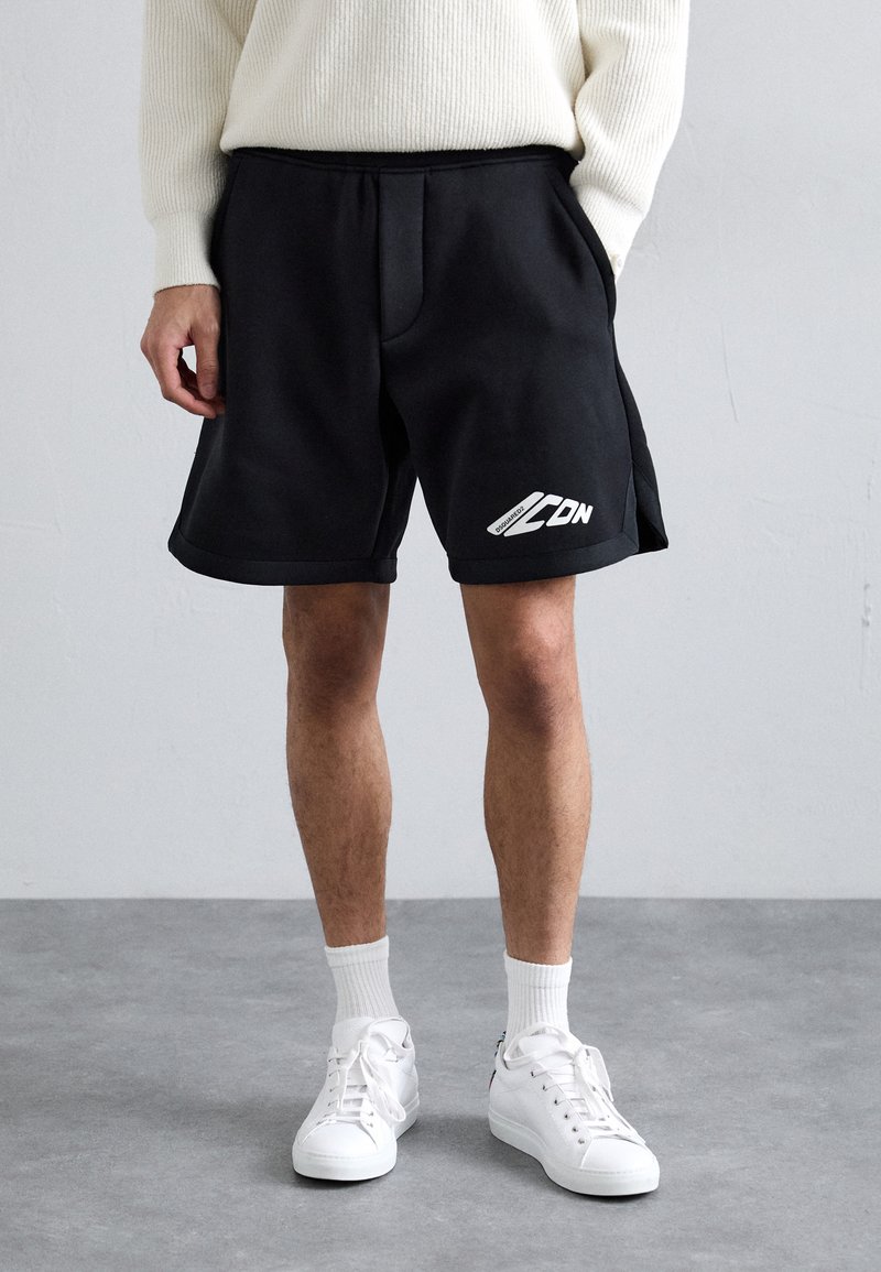 Shorts noirs au style décontracté avec poches latérales et détail logo blanc sur la jambe droite. Portés avec des baskets blanches et des chaussettes blanches côtelées.