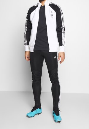 Adidas tepláková souprava s bílou a černou bundou na zip, černé elastické kalhoty a tyrkysové kopačky na fotbal; obsahuje černé detaily a tři pruhy.