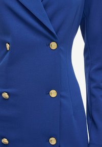 Royal blue blazer dengan pemotongan yang disesuaikan, menampilkan tombol dekoratif emas, desain double-breasted, dan kain yang halus.