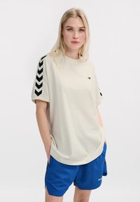T-shirt à manches courtes couleur crème avec des rayures en chevron noires sur les manches, coupe décontractée, et un petit logo sur la poitrine. Associé à un short bleu.