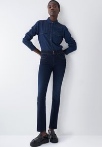 Camisa de mezclilla oscura con cuello abotonado y dos bolsillos en el pecho, combinada con pantalones oscuros de tiro alto y mocasines negros brillantes.