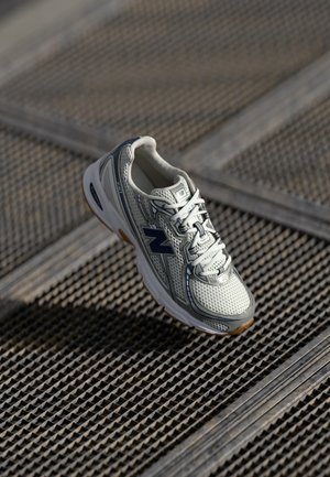 Biało-szary sneaker New Balance ze sznurowadłami, umieszczony na metalowej powierzchni ze stalowej kratki z ukośnymi belkami wspierającymi.
