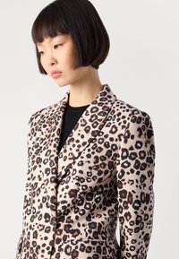 Vrouw met kort donker haar draagt een beige colbert met luipaardprint over een zwarte top en kijkt omlaag tegen een effen lichte achtergrond.