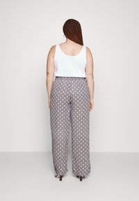 Vrouw met lang vlecht haar, staande, gekleed in een witte mouwloze top, geprinte wijde broek en zwarte hakken, met de rug naar de kijker gekeerd.