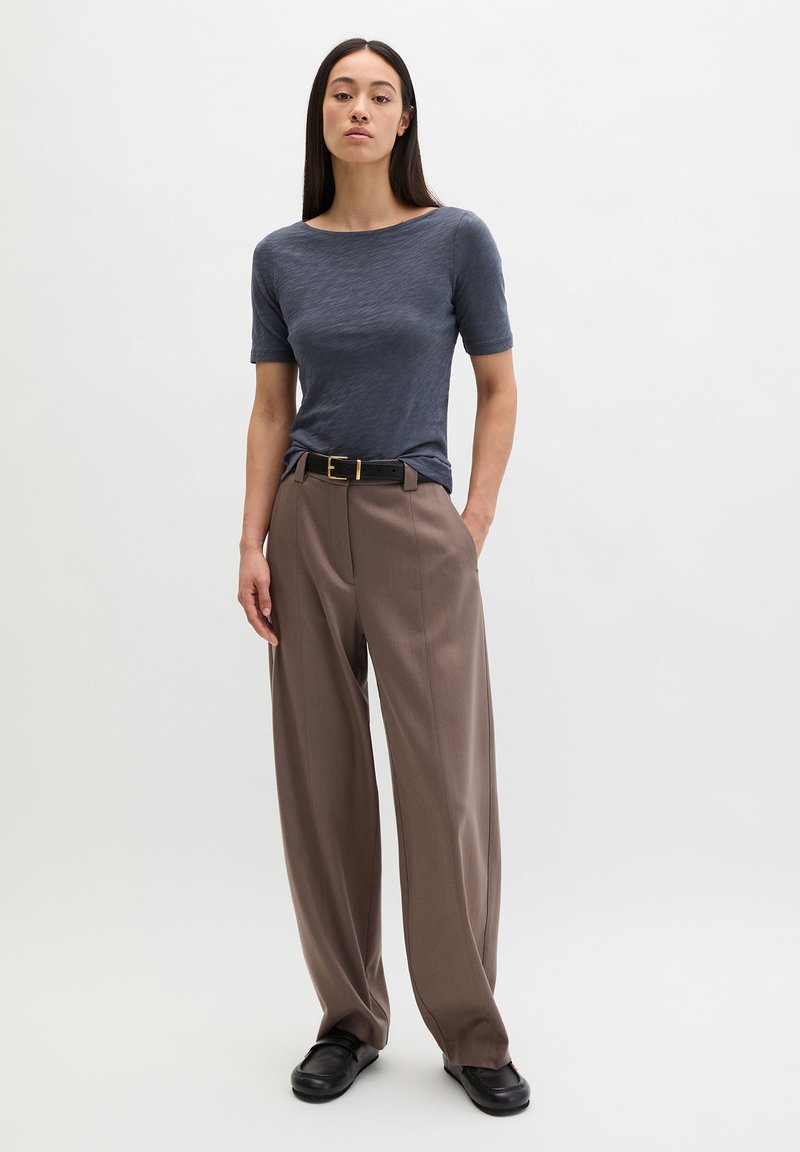 Femme debout portant un haut gris à manches courtes, un pantalon large marron avec une ceinture noire, et des chaussures noires, sur un fond clair uni.