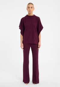 Lila Oberteil mit hohem Kragen und Flügelärmeln, kombiniert mit passenden ausgestellten Hosen. Der Stoff ist glatt mit einem leichten Glanz.