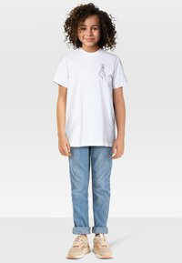 Mala djevojčica s kovrčavom kosom nosi bijelu majicu s dizajnom ruke u obliku srca, svijetloplave traperice povučene na manžetama i bež tenisice.