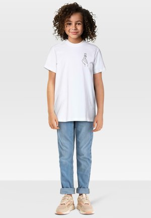 Malá dívka s kudrnatými vlasy, oblečená v bílem tričku s designem srdce z rukou, světle modrých džínách s ohrnutými nohavicemi a béžových teniskách.