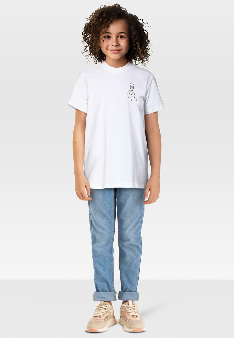 Mala djevojčica s kovrčavom kosom nosi bijelu majicu s dizajnom ruke u obliku srca, svijetloplave traperice povučene na manžetama i bež tenisice.