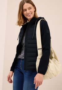 Vadderad svart zip-up jacka med huva, svart och vit randig skjorta under, blåjeans och en beige axelväska.